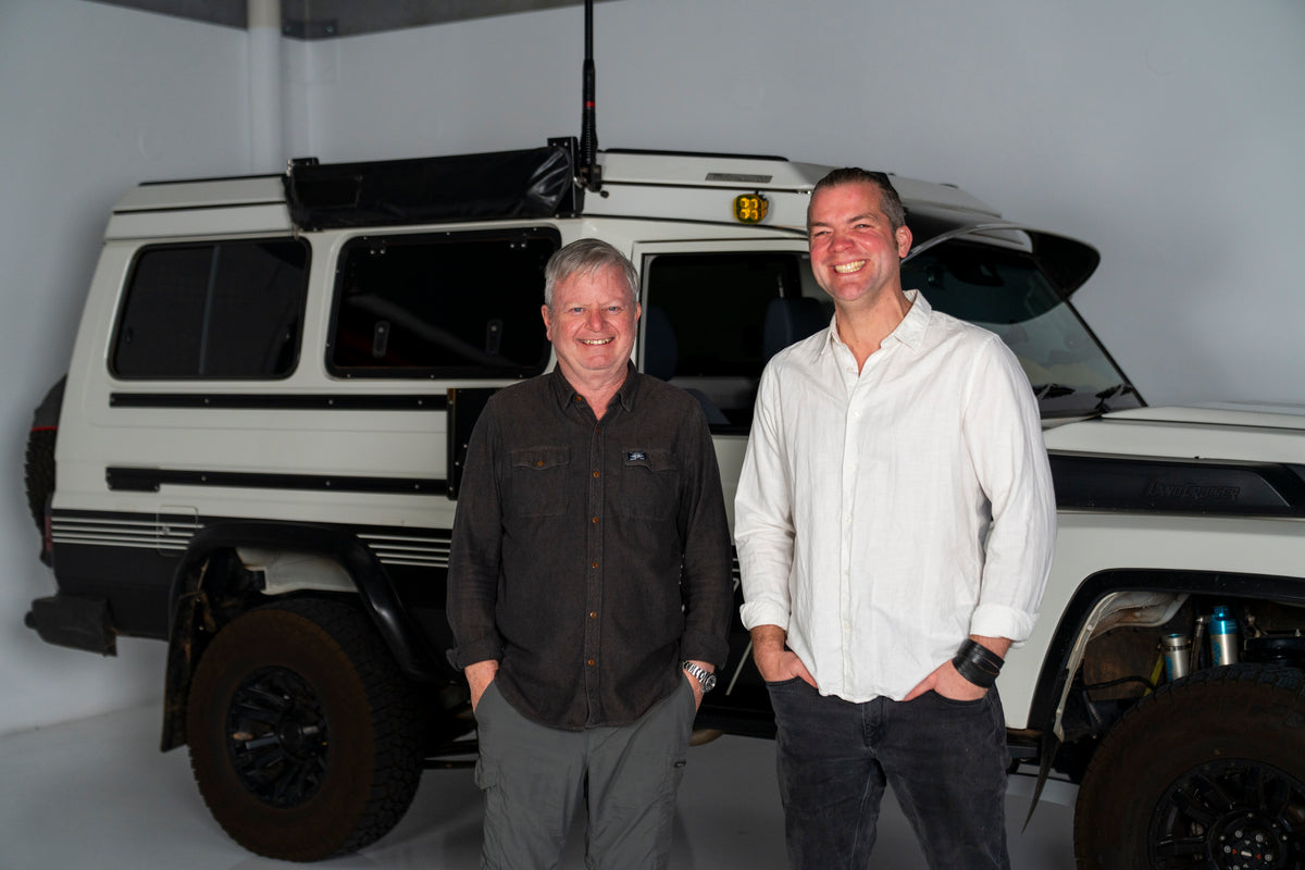 Two men standing in front of a white van with a black roof rack in a garage.